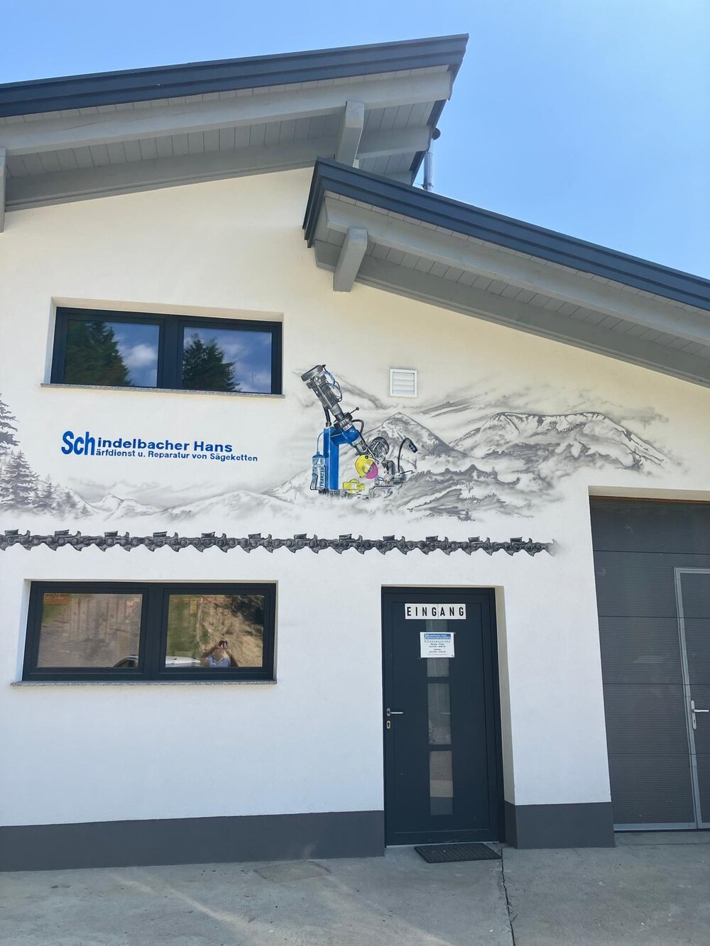 Fassade des Betriebs Schindelbacher Hans mit Wandgemälde einer Gebirgslandschaft und einer blauen Schärfmaschine